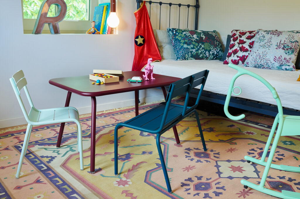 Une salle de jeux avec une table rouge, une chaise enfant colorée de style luxembourgeois, un cheval en bois et un dinosaure rose. Un livre et une boîte en bois ornent le plateau de la table. Des coussins à motifs reposent sur le lit en arrière-plan, ajoutant une touche de charme douillet.