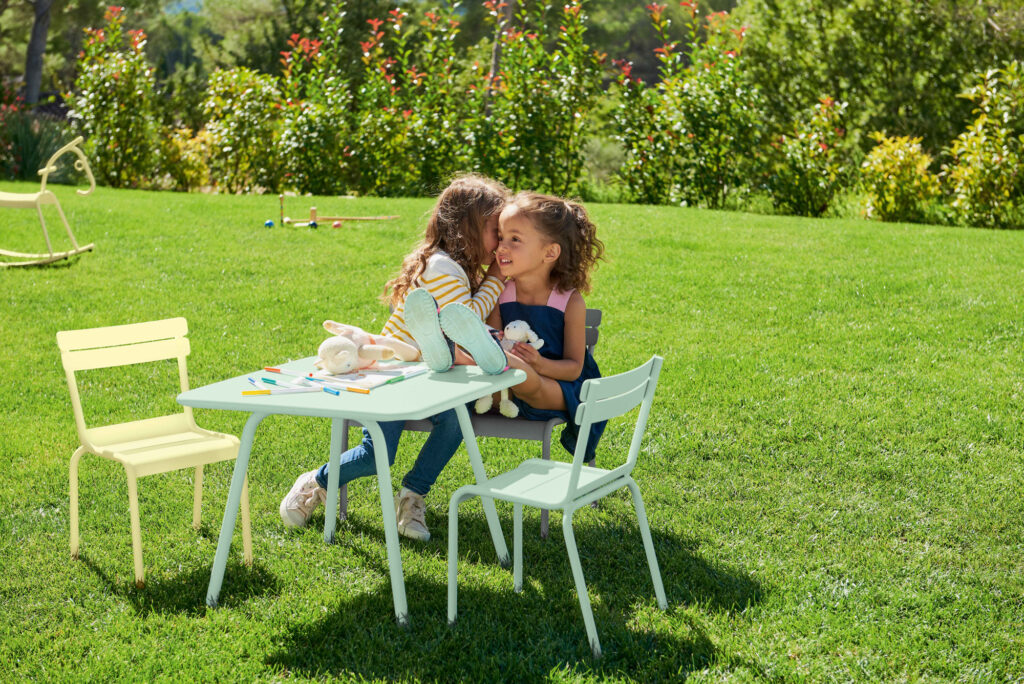 Deux enfants sont assis sur une chaise enfant luxembourgeoise, entourés de jouets et de crayons de couleur, sur une pelouse herbeuse avec un paysage arboré.