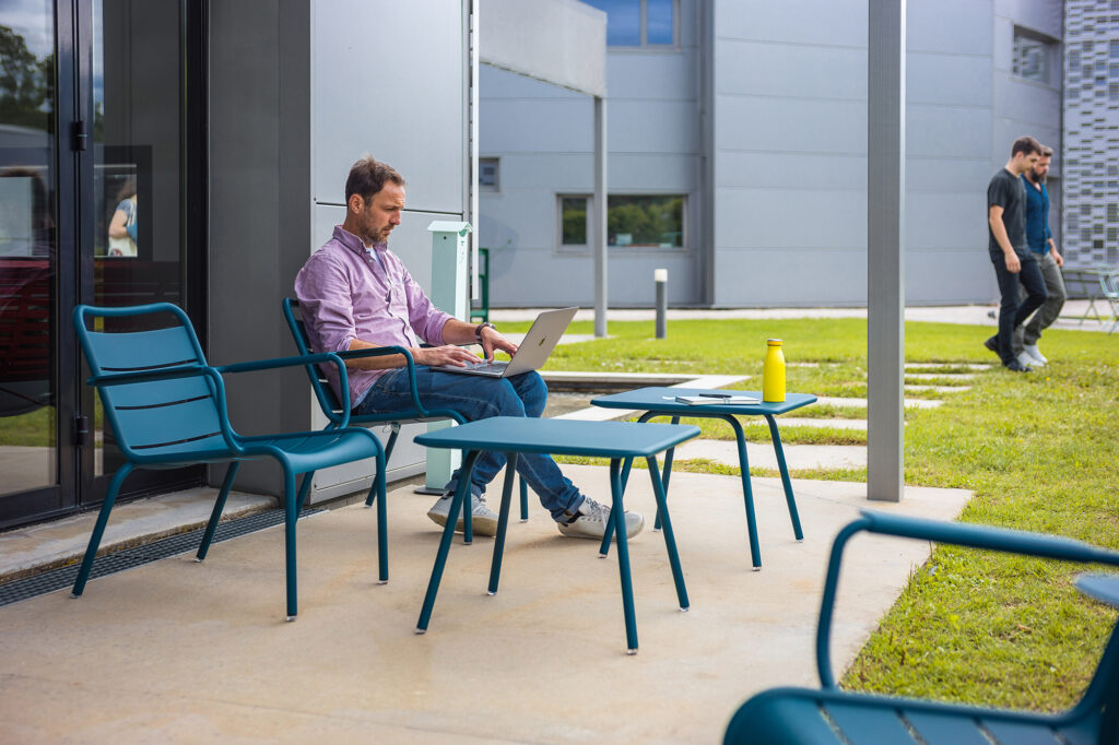 Un homme se détend sur une terrasse, concentré sur son ordinateur portable, assis devant une Table Enfant Luxembourg et des chaises, une bouteille d'eau jaune à ses côtés. Non loin de là, une autre personne déambule, évoquant l'ambiance animée des quartiers animés de Luxembourg.
