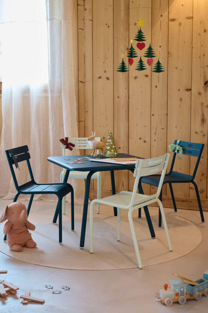 Une charmante table d'enfant ornée de chaises bleues et blanches, d'un petit sapin, de jouets et de pancartes. Cette table enfant Luxembourg est sublimée par un mur en bois et des stickers de Noël en arrière-plan, créant une ambiance chaleureuse et festive.