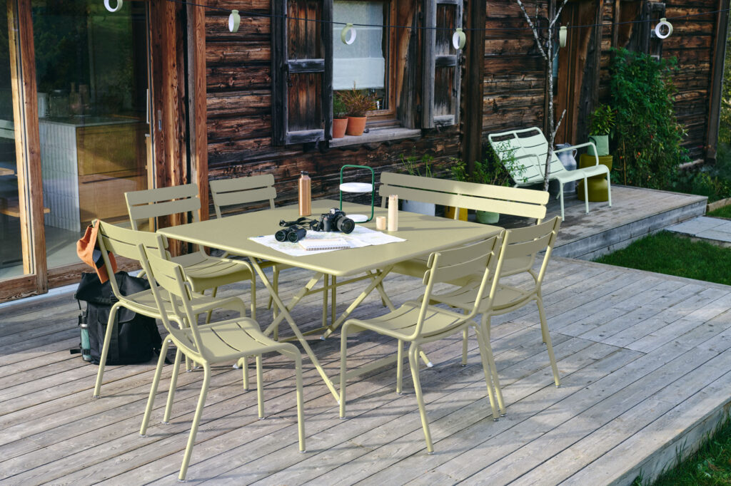 Une terrasse extérieure en bois abrite une table pliante carrée « Caractère » et six chaises vert pâle. Cette table, riche en caractère, est ornée d'une lanterne, d'une carte, de jumelles et d'un carnet. Des guirlandes lumineuses scintillent au-dessus de la terrasse, tandis qu'une charmante maison en bois se niche dans un écrin de verdure à proximité.