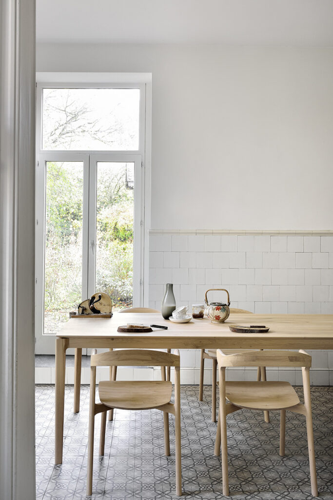 Le coin repas minimaliste comprend la table Bok en chêne, des chaises assorties, un service à thé et un carrelage à motifs, le tout à côté d'une grande fenêtre donnant sur le jardin.