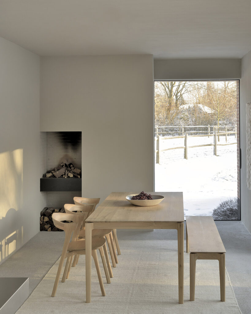 Salle à manger minimaliste avec une table Bok en chêne en bois clair, quatre chaises et un banc, dotée d'une cheminée encastrée avec des bûches empilées, de murs neutres et d'une grande fenêtre révélant une vue extérieure enneigée.