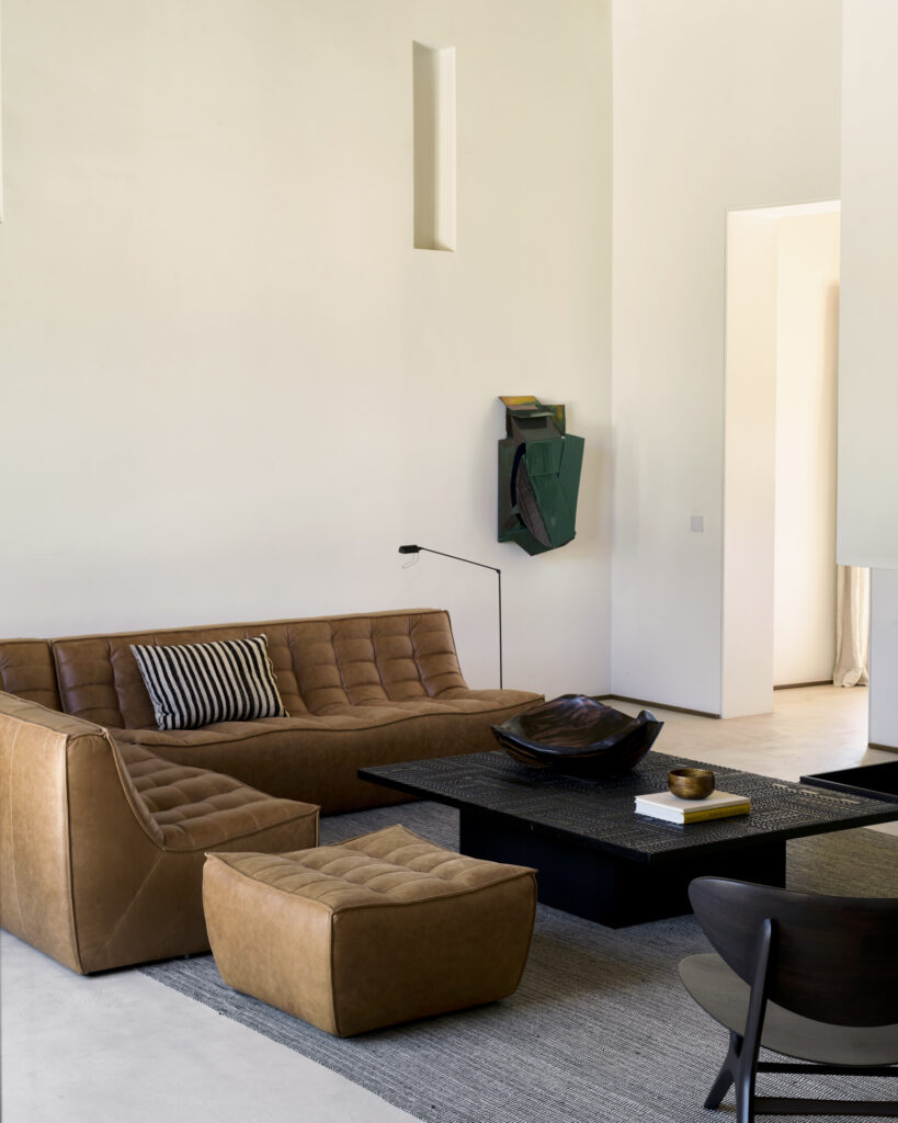 Salon moderne avec un canapé sectionnel N701 en cuir brun, un ottoman Repose-pieds N701, une table basse noire, un tapis neutre et une décoration murale minimaliste. La lumière naturelle éclaire l'espace.