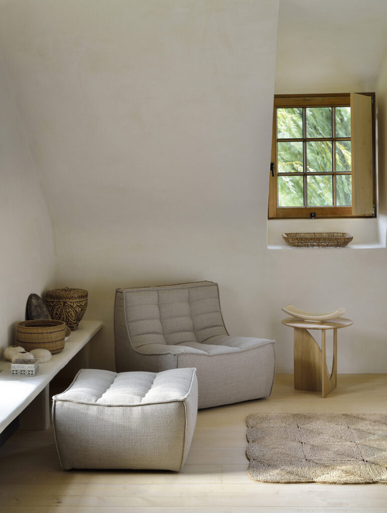 Une pièce minimaliste avec une chaise longue beige N701, des repose-pieds assortis, un tapis tissé, une petite table d'appoint en bois, des paniers décoratifs et une fenêtre ouverte qui laisse entrer la lumière naturelle.