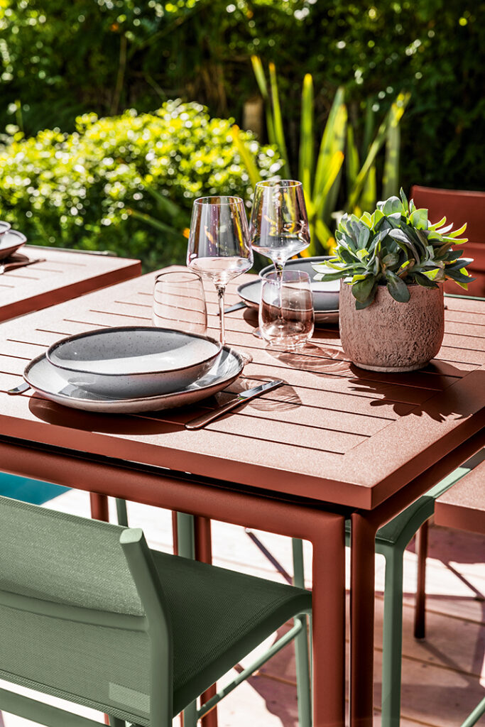 Table carrée Costa avec assiettes, verres à vin et couverts, avec une pièce maîtresse en pot de plantes succulentes ; plantes vertes en arrière-plan.