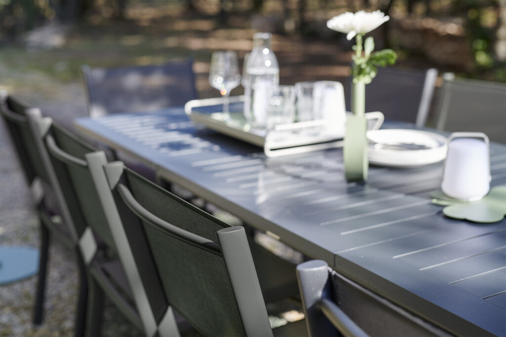 La table extensible Costa est associée à des chaises noires et sertie de verres, d'assiettes, d'une bouteille d'eau et d'une seule fleur blanche dans un vase pour une expérience culinaire en plein air élégante.