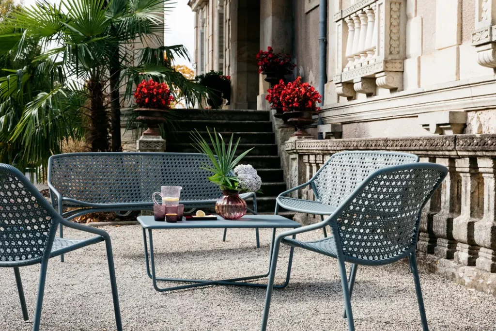 Terrasse extérieure avec Banquette Croisette XL, 3 places, chaises et table en métal, vase à fleurs, pichet et verres, aménagée près d'un bâtiment historique en pierre décoré de pots de fleurs rouges et de palmiers.