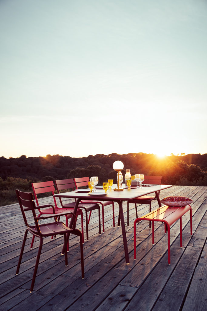 Chaises, table et banc de jardin Luxembourg en aluminium laque dans les tons rouge sur une terrasse avec couché de soleil et verdure