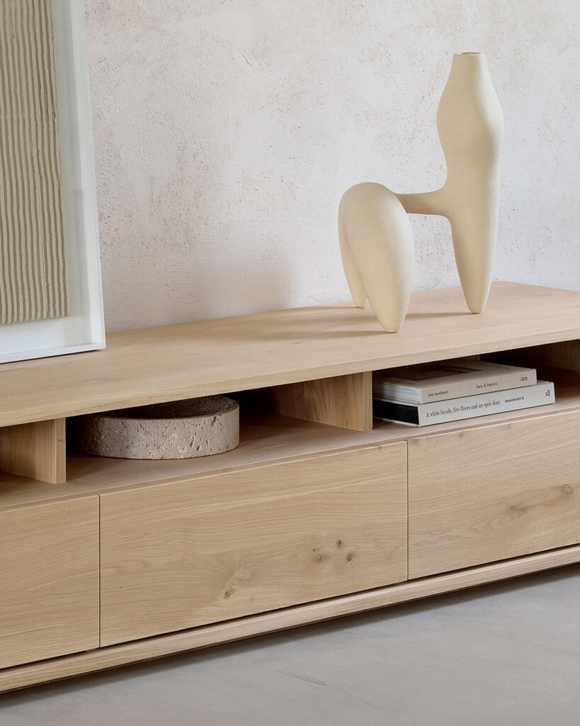 Le Meuble TV Shadow en chêne 3 tiroirs, une console en bois clair, fabriquée à partir d'un élégant chêne, présente une sculpture abstraite, des livres et un objet circulaire. Positionné contre un mur de couleur neutre, il projette une ombre subtile qui ajoute de la profondeur à l'espace.