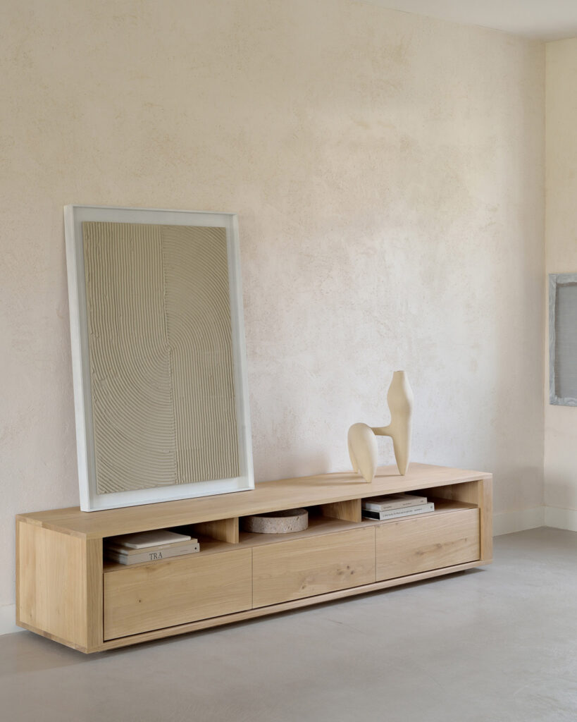 Une pièce minimaliste avec une console basse en bois affichant une œuvre d'art texturée encadrée, une sculpture abstraite et des livres. Les ombres jouent doucement sur le Meuble TV Shadow en chêne 3 tiroirs, créant une esthétique accueillante.