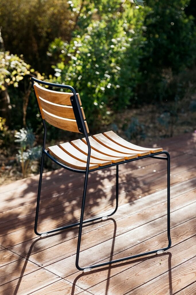 Une Chaise Surprising à lattes en bois de teck avec une structure en métal noir sur une terrasse en teck, baignée de soleil, entourée de verdure.