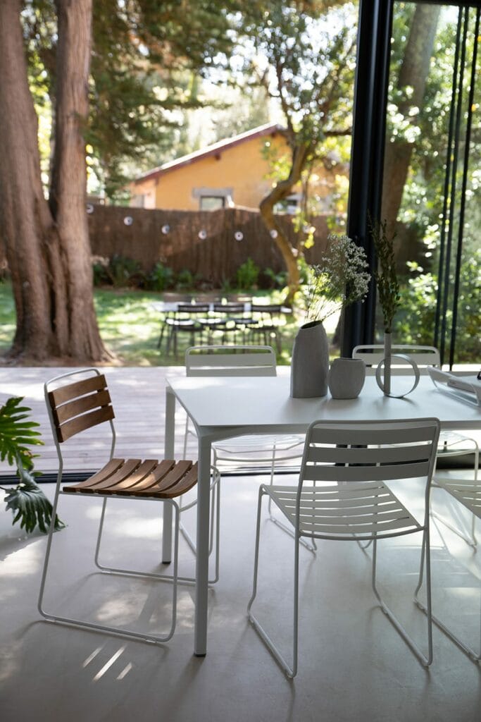 Patio extérieur moderne avec chaise élégante et mobilier en teck surprenant, donnant sur un jardin luxuriant, vu à travers des baies vitrées.