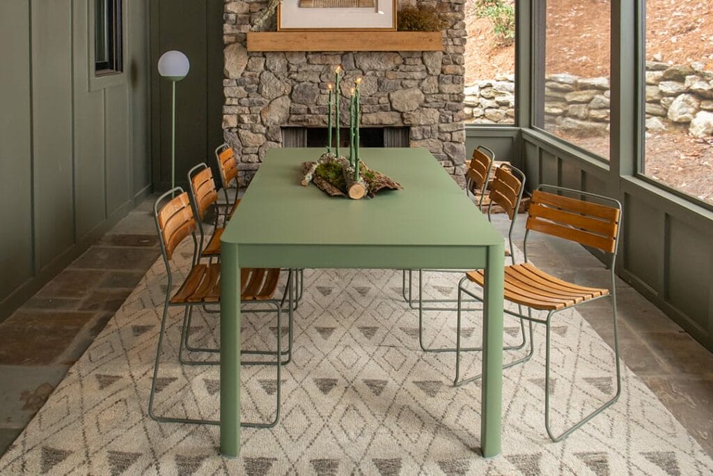 Un coin repas rustique avec une table verte, une chaise Surprising en teck et une cheminée en pierre, donnant sur une vue extérieure à travers de grandes fenêtres.