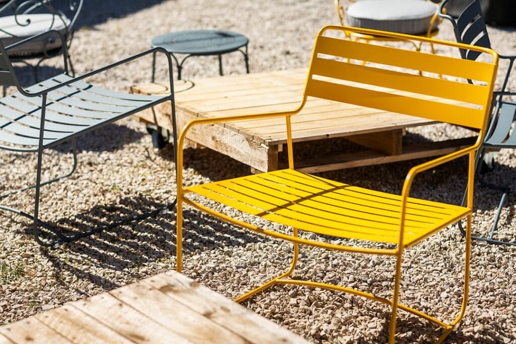 Coin salon extérieur avec une chaise jaune Eames Aluminium Group et un banc en bois sur du gravier, entouré d'autres chaises et tables sous la lumière du soleil.