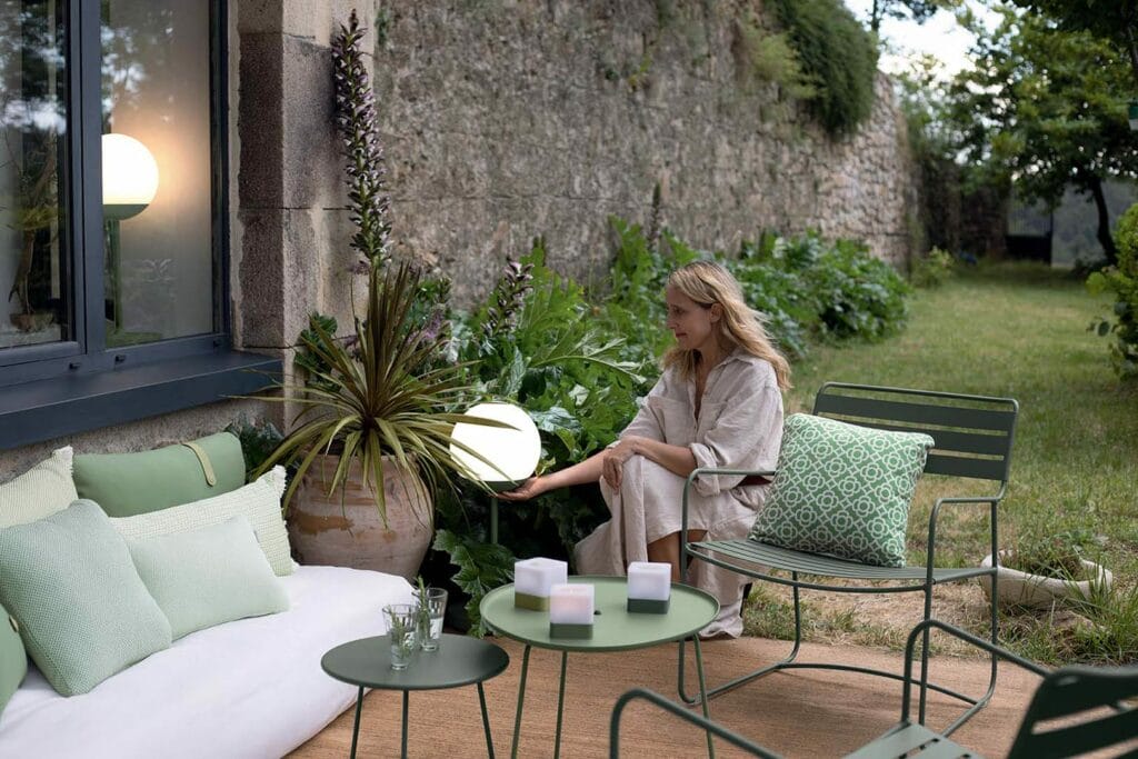 Une femme assise sur un coussin de terrasse près d'un mur de pierre, ajustant une lampe sphérique à côté du Fauteuil bas vert. Meubles et plantes surprenants.