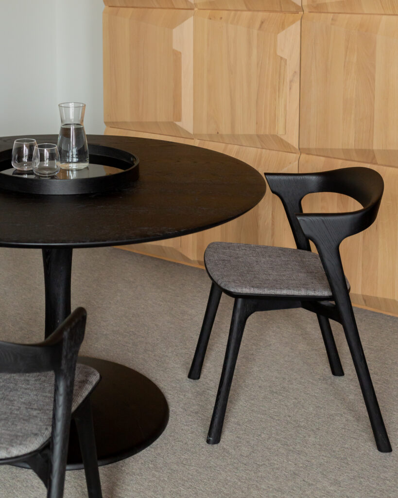 Une table ronde noire avec une carafe et des verres dessus, deux chaises Chaise Bok en chêne noir avec coussins en tissu gris et des armoires en bois en arrière-plan.