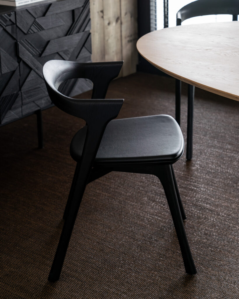Une chaise Bok en chêne noir, assise en cuir avec un dossier incurvé est placée à côté d'une table ronde en bois sur un sol en moquette foncée.