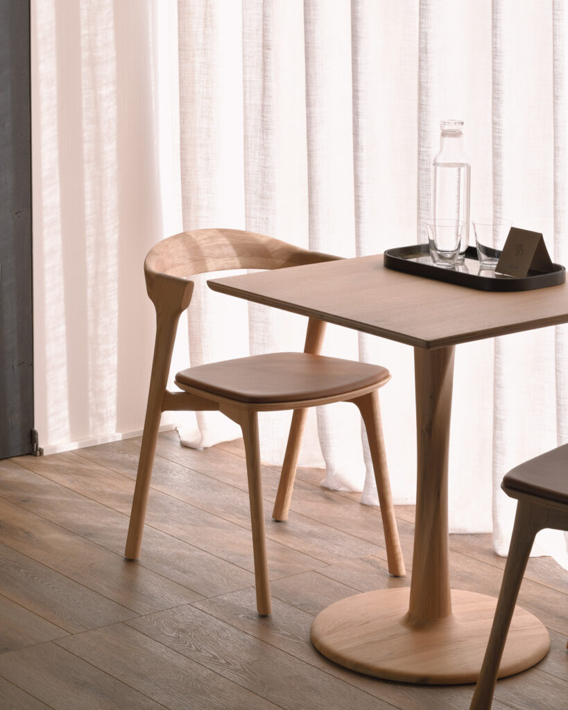 Une chaise en bois minimaliste et une table ronde sont placées près de rideaux blancs transparents. La table est composée d'un plateau noir, d'une bouteille en verre d'eau et d'un verre à l'envers. La finition Chaise Bok en chêne, assise en cuir Cognac du meuble ajoute une touche d'élégance à ce cadre serein.