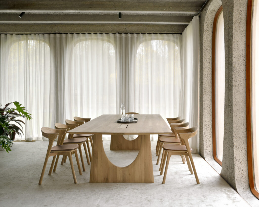 Une salle à manger moderne dispose d'une table en bois de chêne avec huit chaises Bok en chêne, assise en cuir Cognac, entourées de grandes fenêtres avec des rideaux transparents tirés. Il y a une plante sur le côté gauche de la table.