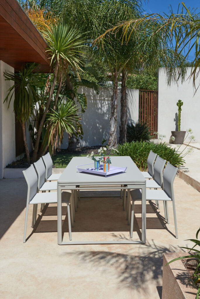 Le patio extérieur présente un ensemble de salle à manger extensible Biarritz gris chic avec six chaises élégantes, orné d'une petite pièce maîtresse et rehaussé de palmiers et de plantes ajoutant une beauté naturelle à l'espace ensoleillé.