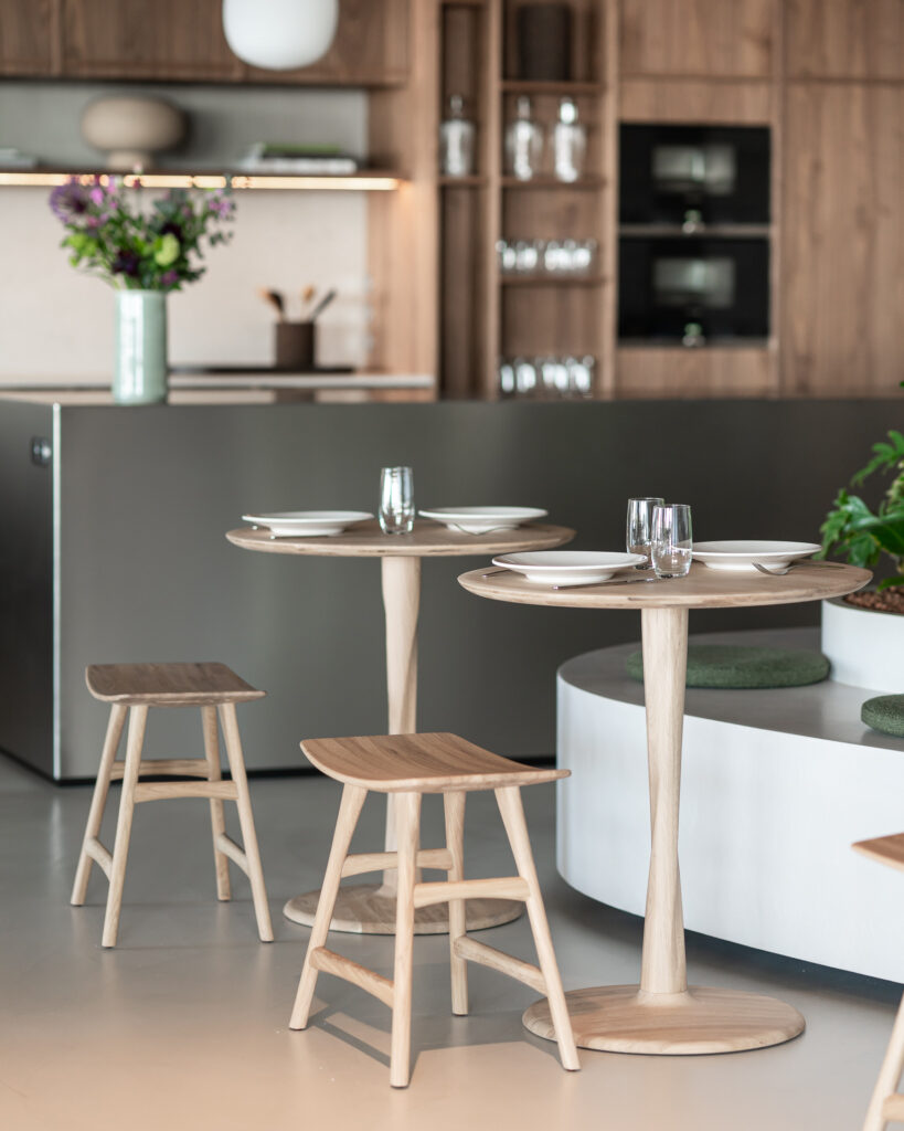 Deux tables Torsion en chêne et leurs tabourets assortis sont disposés avec des assiettes et des verres dans une salle à manger moderne et minimaliste, dotée d'armoires en bois et de plantes décoratives, ajoutant une touche unique de raffinement à l'espace.