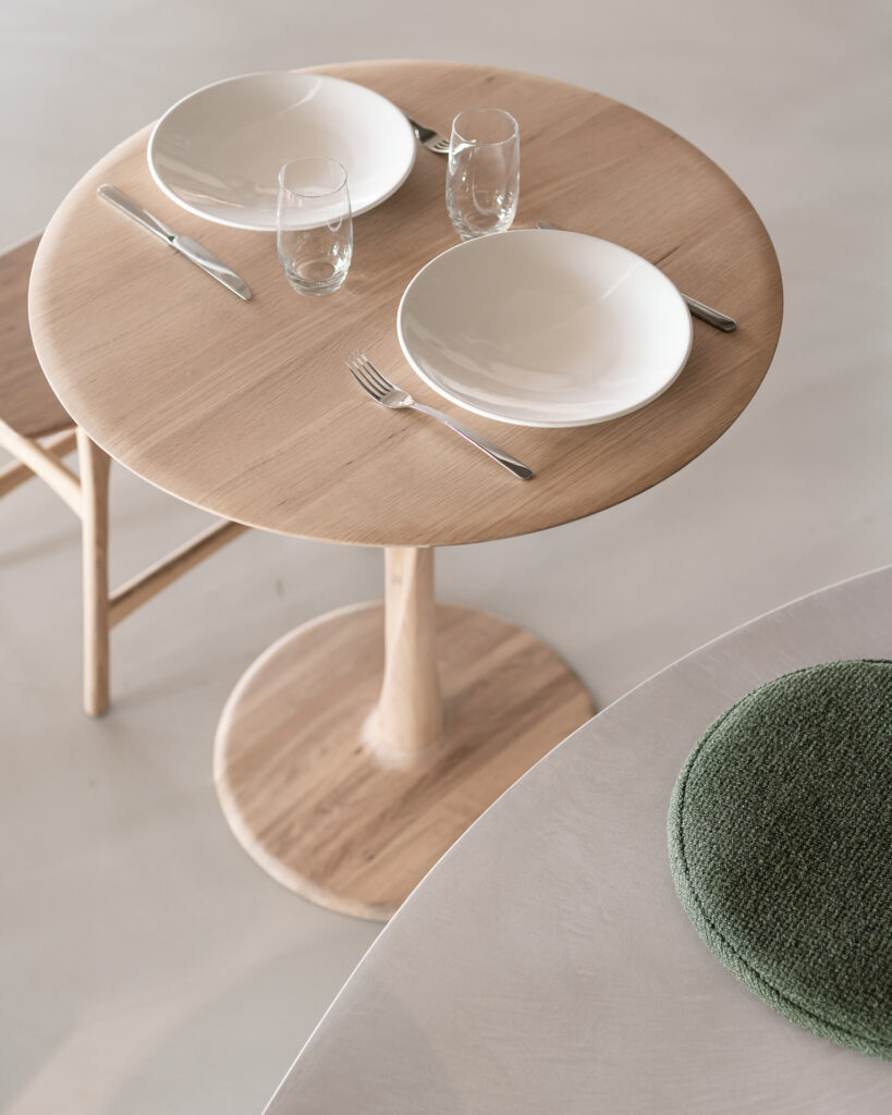 Un ensemble Table Torsion en chêne pour deux personnes comprend des assiettes blanches, des fourchettes, des couteaux et des verres à eau ; à proximité se trouvent une chaise en bois et un coussin vert.