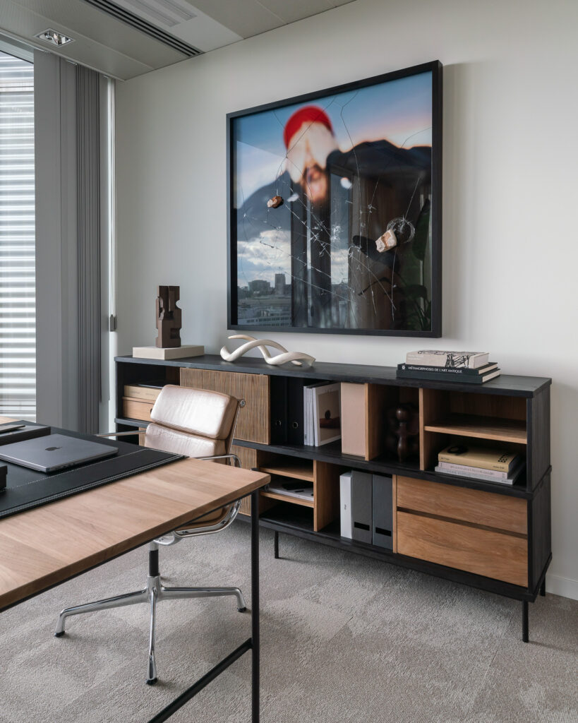 Espace bureau avec un buffet en teck Oscar faisant office de bureau et de chaise, un meuble de rangement en bois avec étagères et classeurs, et une grande œuvre d'art encadrée au mur.