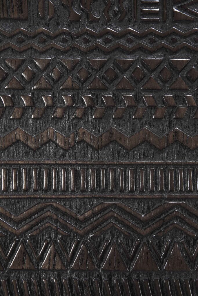 Gros plan d'une surface texturée aux motifs géométriques complexes sculptés dans le bois foncé du Buffet Tabwa en teck noir 3 portes, reflétant l'esthétique du teck noir.