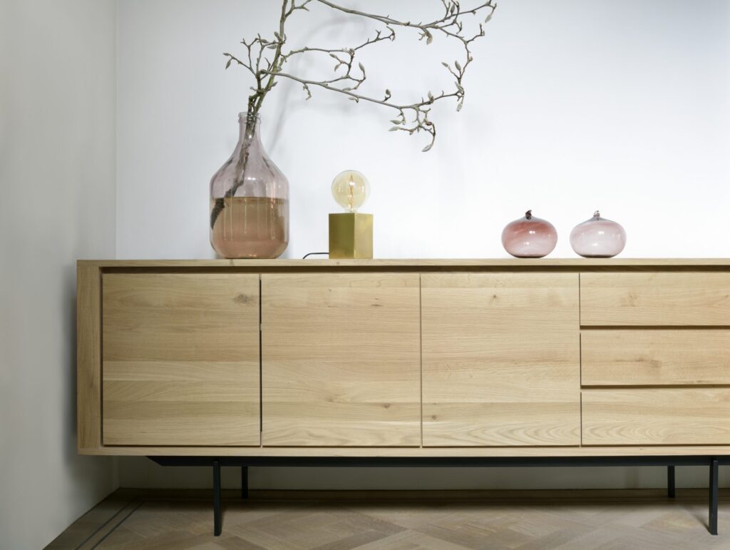 Le Buffet Shadow, chêne, 3 portes, 3 tiroirs, présente un design épuré et minimaliste avec une finition en bois. Il est orné d'éléments décoratifs dont un vase en verre, une lampe et des objets décoratifs roses sur le dessus, ainsi qu'une branche s'étendant élégamment du vase.