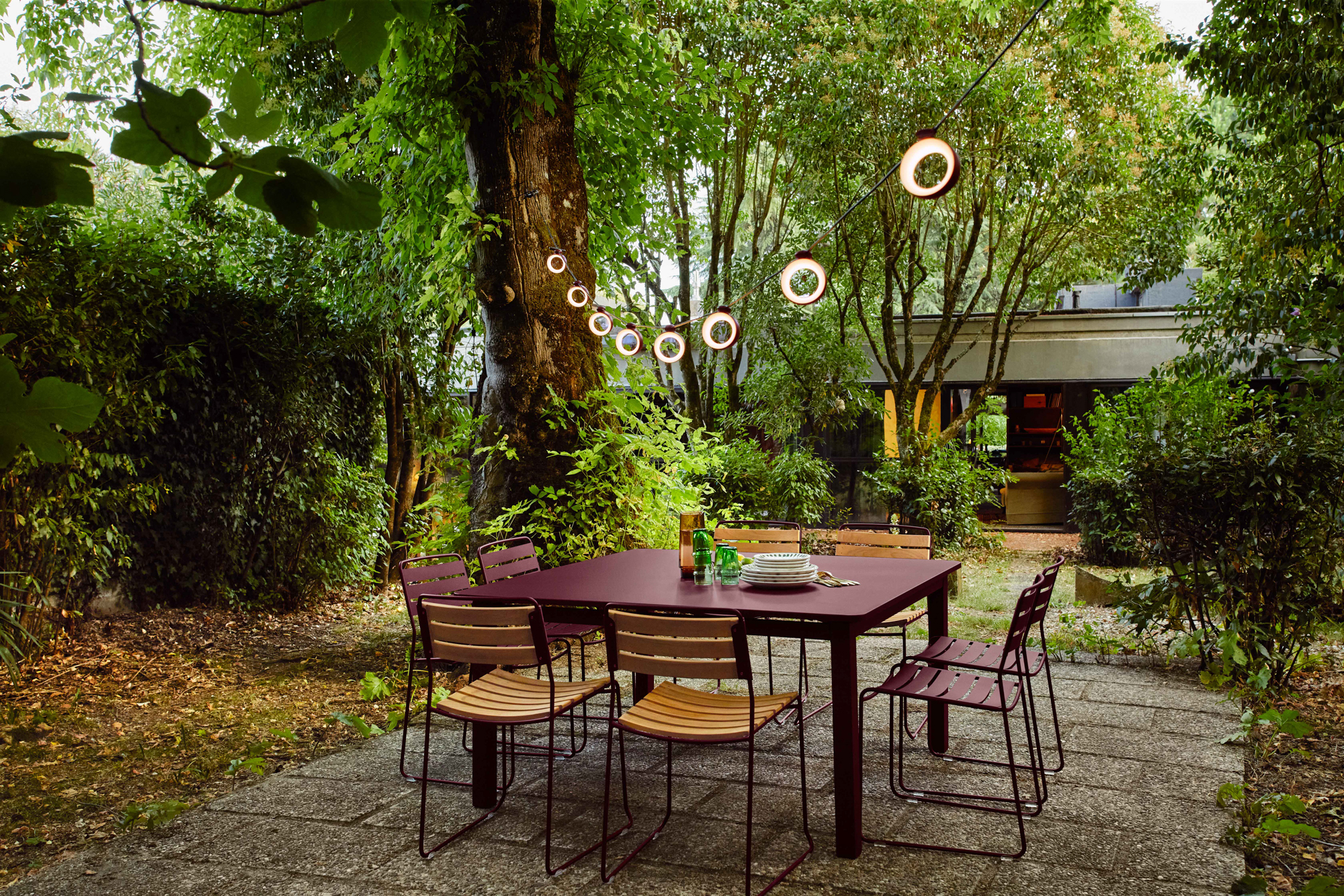 Préparez le décor de votre Table Craft avec une charmante salle à manger extérieure comprenant une table marron, six chaises et de charmantes guirlandes lumineuses suspendues entre les arbres dans un jardin verdoyant et luxuriant.