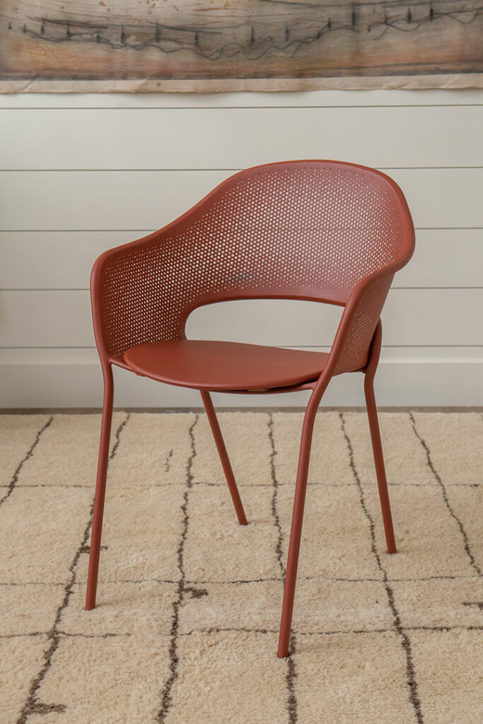 Le Fauteuil Kate, une chaise rouge moderne avec un dossier et une assise perforés, positionnée sur un tapis beige avec un mur beige et gris en arrière-plan, ajoute une touche de style à l'espace de vie contemporain de Kate.