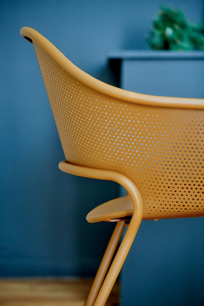 Vue rapprochée du Fauteuil Kate, une chaise moderne en plastique perforé orange, contre un mur bleu-gris.