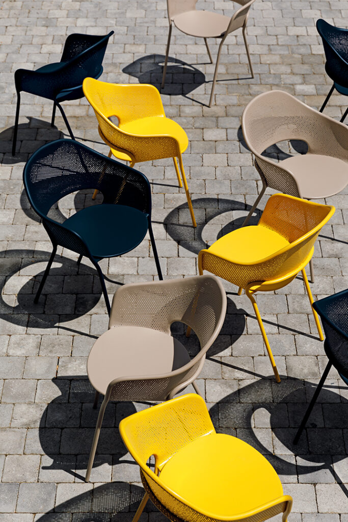 Un groupe de chaises Fauteuil Kate en bleu, jaune et beige sont disposés à l'extérieur sur une surface pavée, projetant des ombres distinctes ; chaque Fauteuil Kate est élégant et accueillant sous le soleil.