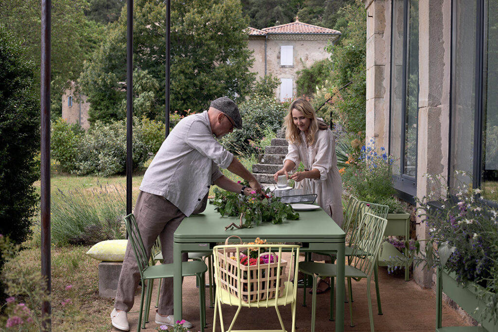 Deux personnes, un homme et une femme, disposent des fleurs et des légumes sur une table verte dans un jardin-terrasse entouré d'une verdure luxuriante, juste à côté d'une chaise Kintbury au design élégant, près du bâtiment en pierre.