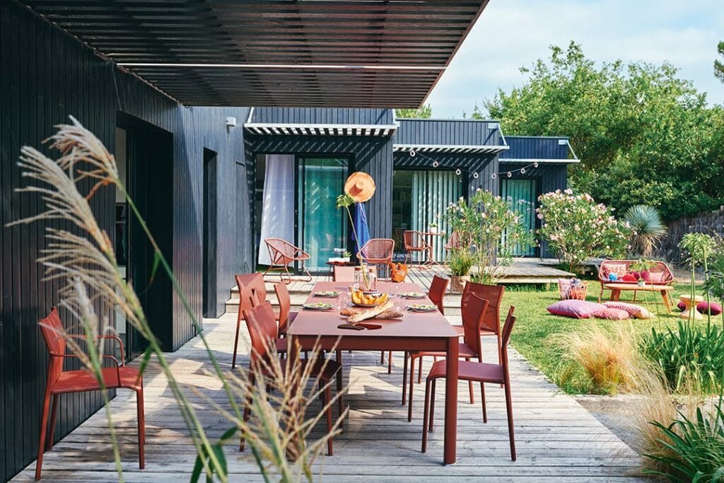 Coin repas extérieur moderne avec une table rouge Chaise Cadiz dressée pour un repas, entouré d'un jardin luxuriant et d'un extérieur de maison noire contemporaine.