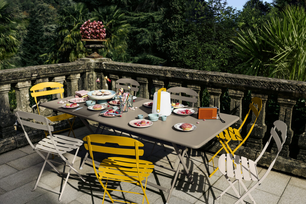 Une table à manger extérieure sur un balcon avec huit chaises est joliment dressée avec des assiettes, des verres, une lanterne et une petite radio, le tout entouré d'une verdure luxuriante. Pour compléter le cadre serein, les chaises Chaise La Môme ajoutent une touche d'élégance.