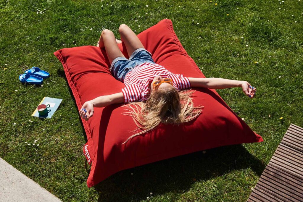 Une jeune fille vêtue d'une chemise à rayures rouges se détend sur un coussin Fatboy Original Outdoor sur l'herbe, les bras tendus. Des sandales bleues et un livre sont posés à proximité, créant une scène extérieure parfaite.