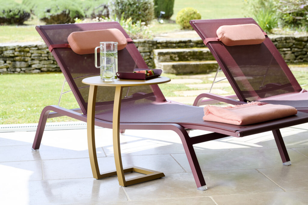 Deux chaises longues avec des coussins roses sont posées sur une terrasse donnant sur un jardin herbeux, à côté d'une table basse déportée Alizé tenant un pichet d'eau, un verre et un plateau.