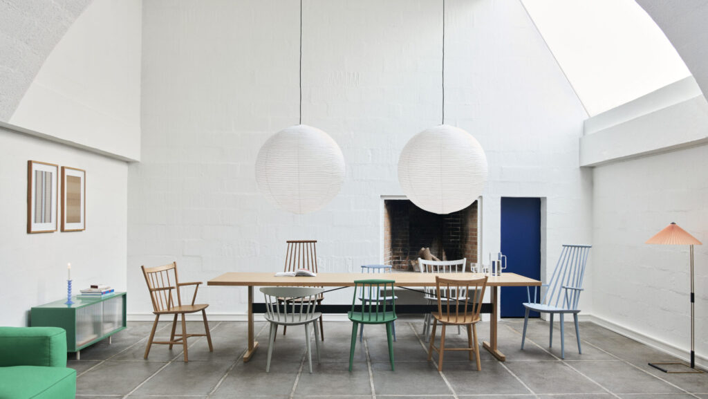 Salle à manger minimaliste avec une table en bois, des chaises dépareillées comme la Stoel J110, deux lampes suspendues rondes, une cheminée, une lucarne, des murs blancs et un sol en dalles de pierre.