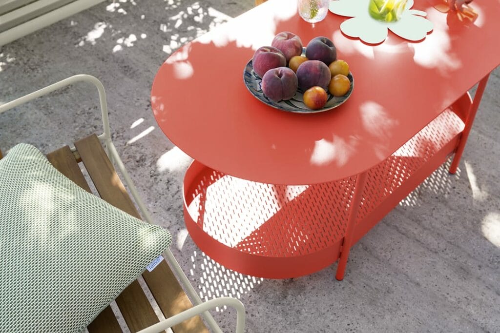 Phrase avec le nom du produit : Un balcon lumineux et ensoleillé avec une table basse Salsa ovale rouge tenant un bol de pêches, à côté d'une chaise pliante avec un coussin rayé.