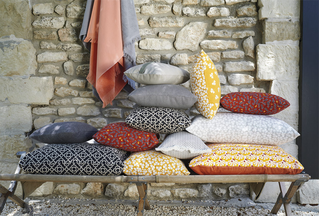 Une pile d'oreillers Coussin Lorette 44x44cm sur un banc devant un mur de pierre.