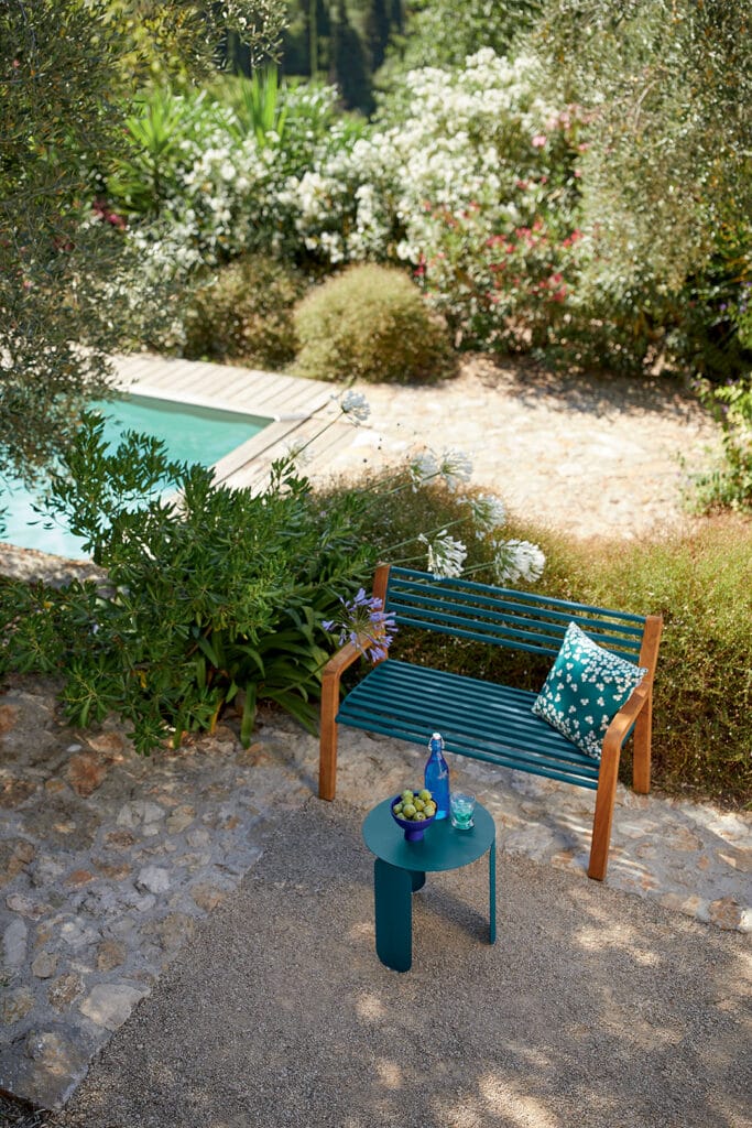 Une scène de jardin tranquille avec une chaise Adirondack à côté d'une petite table contenant un livre et des boissons, donnant sur une piscine entourée de buissons fleuris luxuriants.