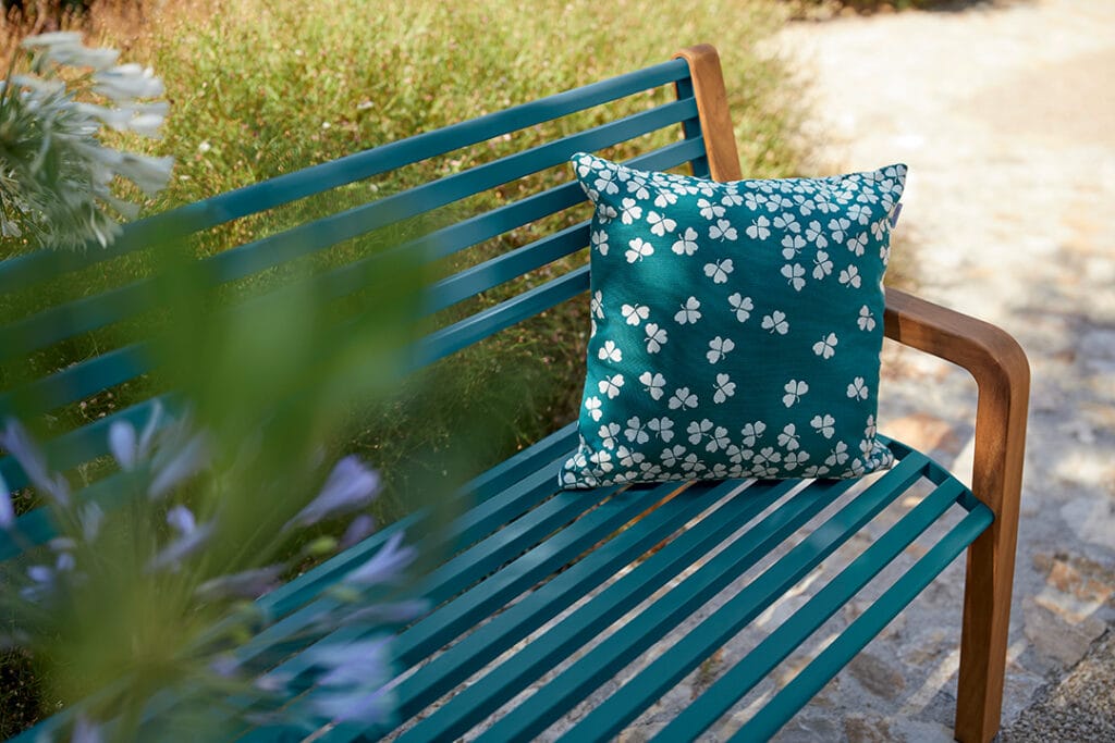 Phrase avec le nom du produit : Un oreiller à motifs floraux bleus et blancs repose sur un « canapé Marlowe » bleu rayé assorti dans un jardin tranquille.