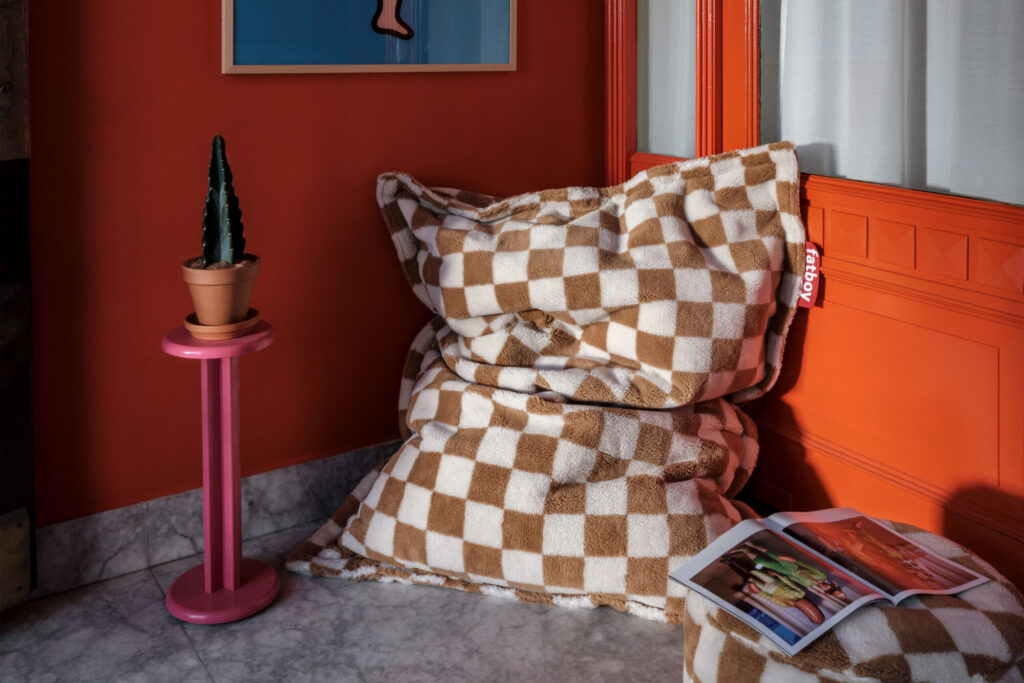 Un pouf Fatboy Original Slim Teddy à carreaux bruns et blancs trône près d'un mur et d'une porte orange, à côté d'une table d'appoint rose contenant un cactus en pot et un magazine ouvert, le tout sur un sol en marbre.