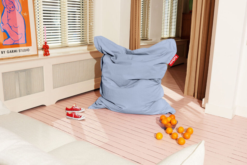 Un grand pouf bleu clair Fatboy Original Stonewashed est posé sur un sol en bois rose à côté de balles orange et de baskets rouges éparpillées, avec des œuvres d'art encadrées et des fenêtres en arrière-plan.