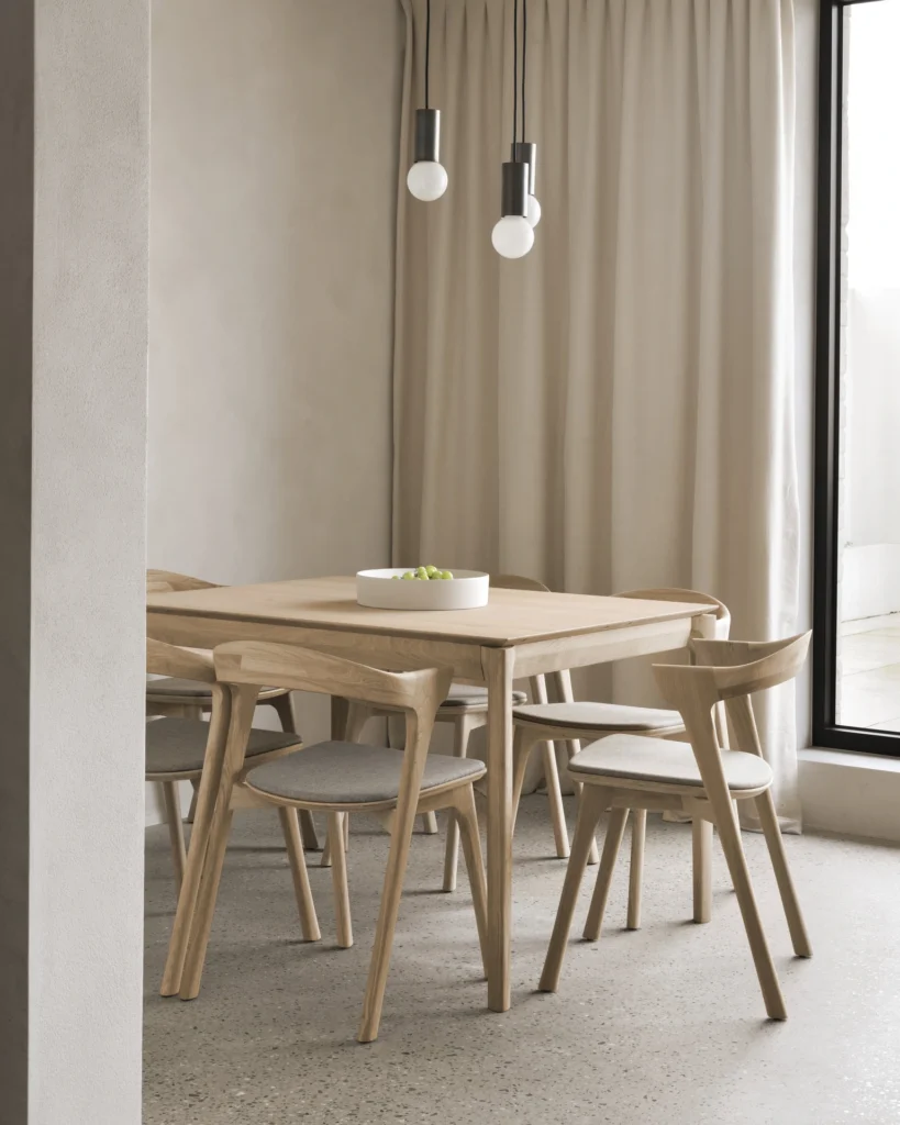 Un coin repas minimaliste avec une Table Bok à rallonge en chêne, quatre chaises assorties, un bol blanc, des lampes suspendues, des rideaux beiges et une grande fenêtre.
