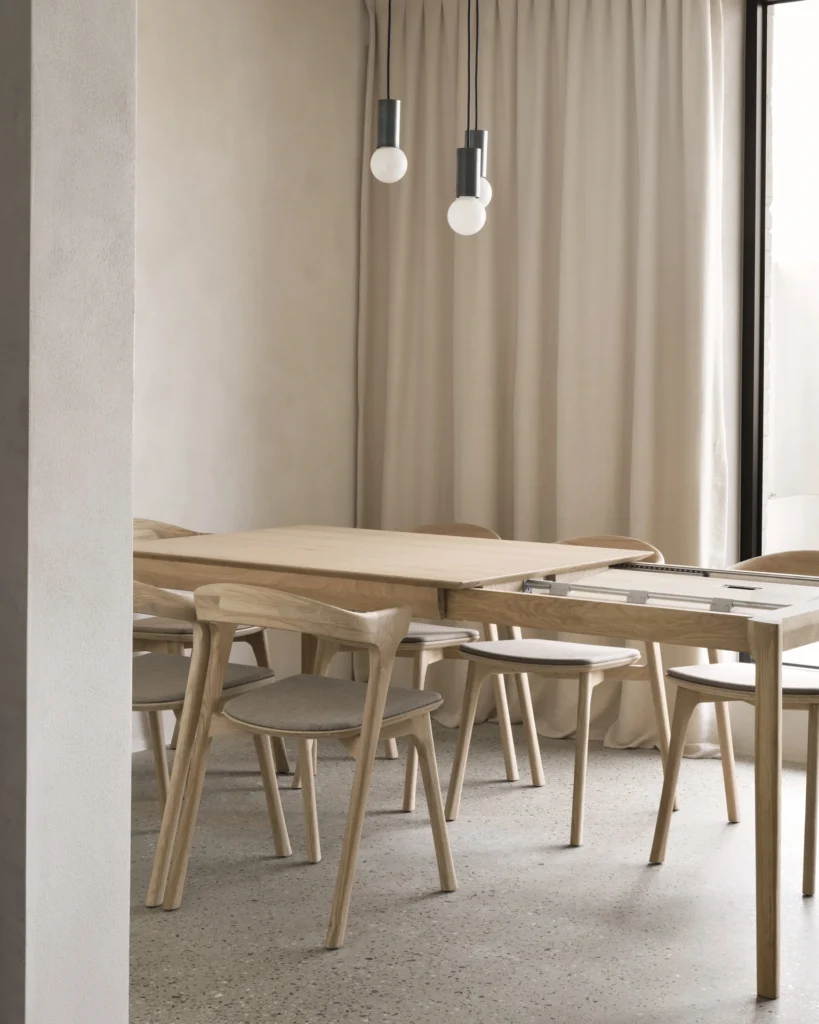 Salle à manger minimaliste avec une Table Bok à rallonge en chêne, des chaises assorties, des rideaux beiges et deux ampoules suspendues au plafond.