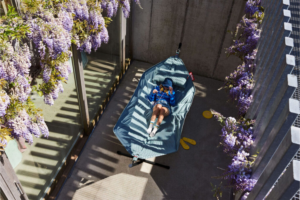Personne en sweat à capuche bleu se relaxant sur un Headdemock Superbe hamac dans une cour en béton, entouré de glycines violettes et de la lumière du soleil. Des pantoufles jaunes sont posées sur le sol à proximité.