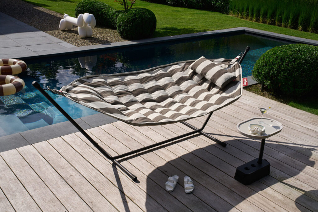 Un Headdemock Superbe hamac d'extérieur avec des rayures et un oreiller repose sur une terrasse en bois au bord de la piscine, à côté de sandales et d'une petite table avec une boisson, le tout entouré d'une verdure luxuriante.